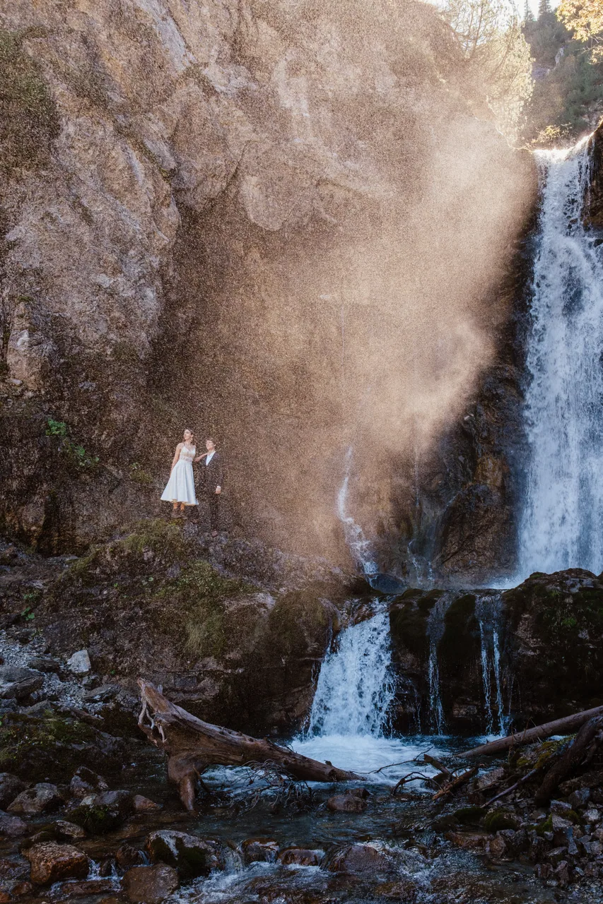 A couple tiny at the foot of a vast alpine waterfall