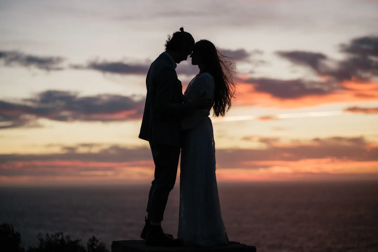 Silhouette of a couple holding hands on a sea cliff at sunset during their elopement ceremony
