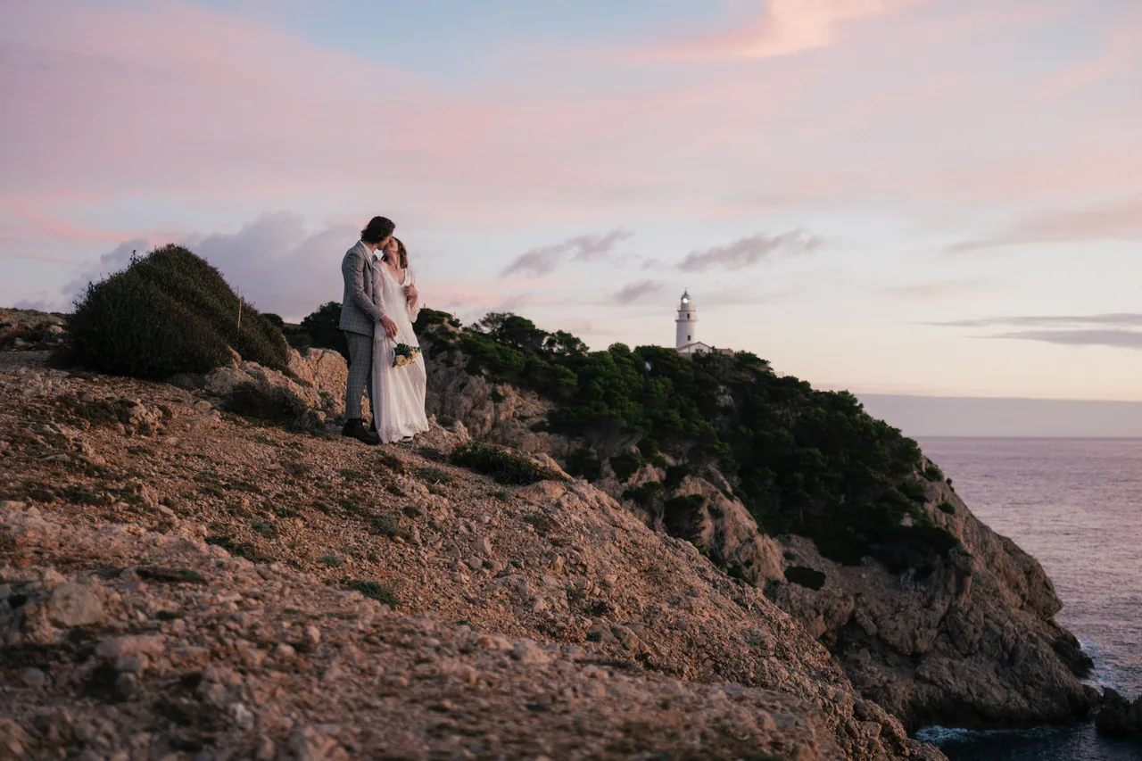 Ein Paar auf einer Klippe über dem Meer mit einem fernen Leuchtturm in der Dämmerung auf Mallorca