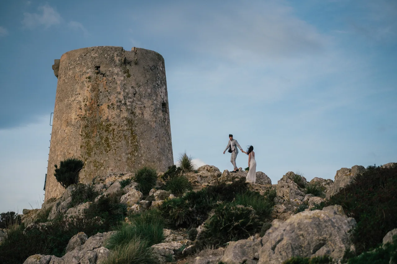 Ein Paar geht über die Küstenfelsen unterhalb eines alten Wachturms auf Mallorca